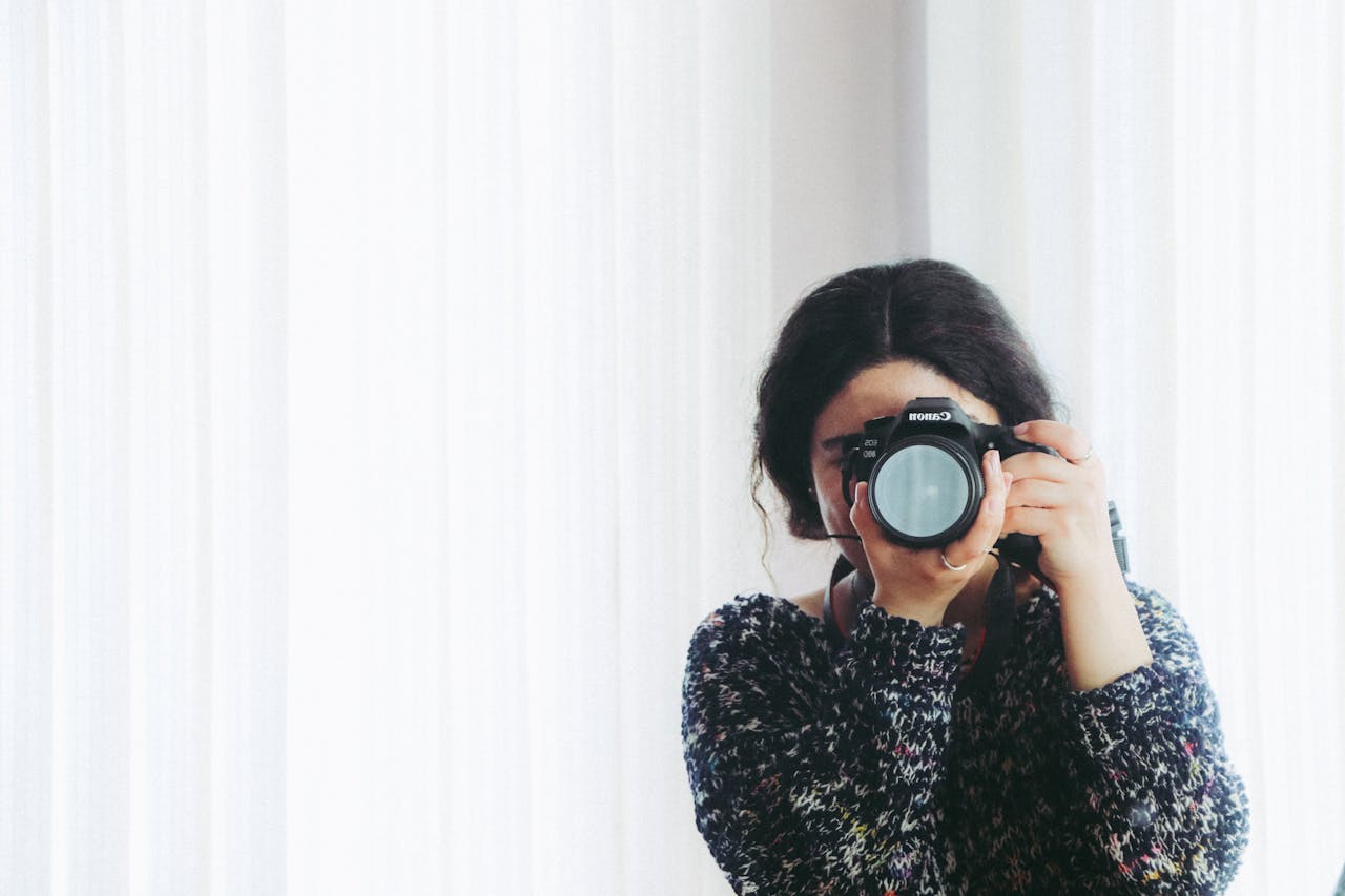 A young woman holding a camera indoors, capturing a photo in a modern setting.